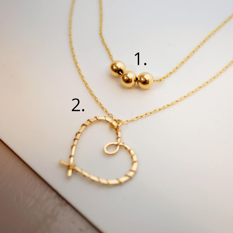 May include: Two gold necklaces. The first necklace has three gold beads. The second necklace has a heart-shaped pendant.