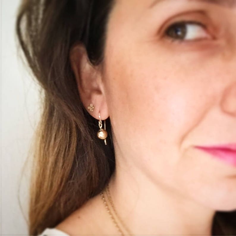 May include: A close-up of a person's ear wearing a pair of gold dangle earrings with a small round ball at the end. The earrings are simple and elegant.