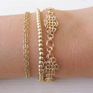 May include: Three gold bracelets stacked on a wrist. The top bracelet is a delicate chain with a small, intricate design. The middle bracelet is a simple chain of small, round beads. The bottom bracelet is a thicker chain with a larger, more elaborate design.