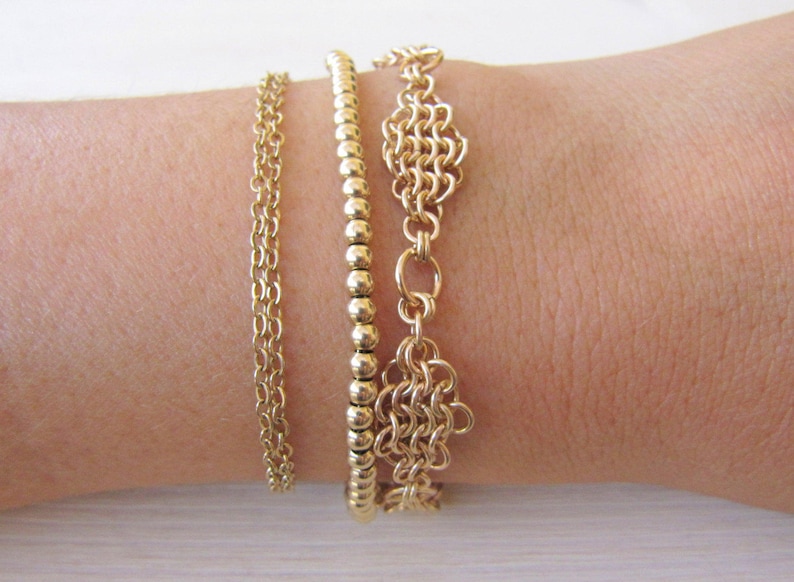May include: Three gold bracelets stacked on a wrist. The top bracelet is a delicate chain with a small, intricate design. The middle bracelet is a simple chain of small, round beads. The bottom bracelet is a thicker chain with a larger, more elaborate design.