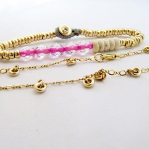 May include: Three gold-toned bracelets are displayed against a white background. One bracelet is made of small, round beads, another features pink and clear beads, and the third is a delicate chain with small, decorative charms.
