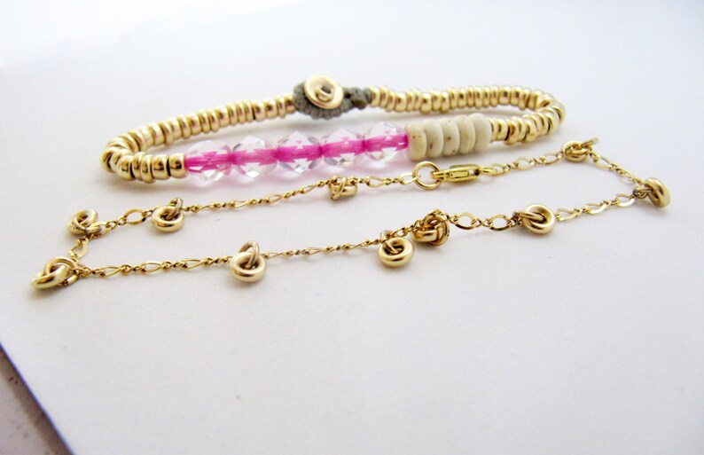 May include: Three gold-toned bracelets are displayed against a white background. One bracelet is made of small, round beads, another features pink and clear beads, and the third is a delicate chain with small, decorative charms.