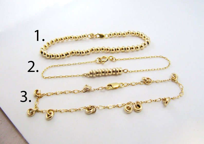 May include: Three gold bracelets with different styles. The first bracelet is a simple chain of gold beads. The second bracelet is a delicate chain with a row of small gold rings. The third bracelet is a chain with small gold rings and a few dangling gold rings.