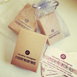 May include: Three brown cardboard boxes with the text "Jewelry Designer" and a small heart symbol printed on them. The boxes are wrapped in a white and gray sheer fabric. The boxes are sitting on a white surface. There are also several business cards with the same text and a website address.