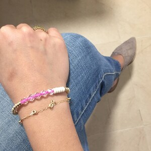 May include: Two bracelets are displayed on a person's wrist. One is a gold chain bracelet with small charms. The other is a beaded bracelet with pink beads and white cylindrical beads, with gold accents. The person is wearing blue jeans.