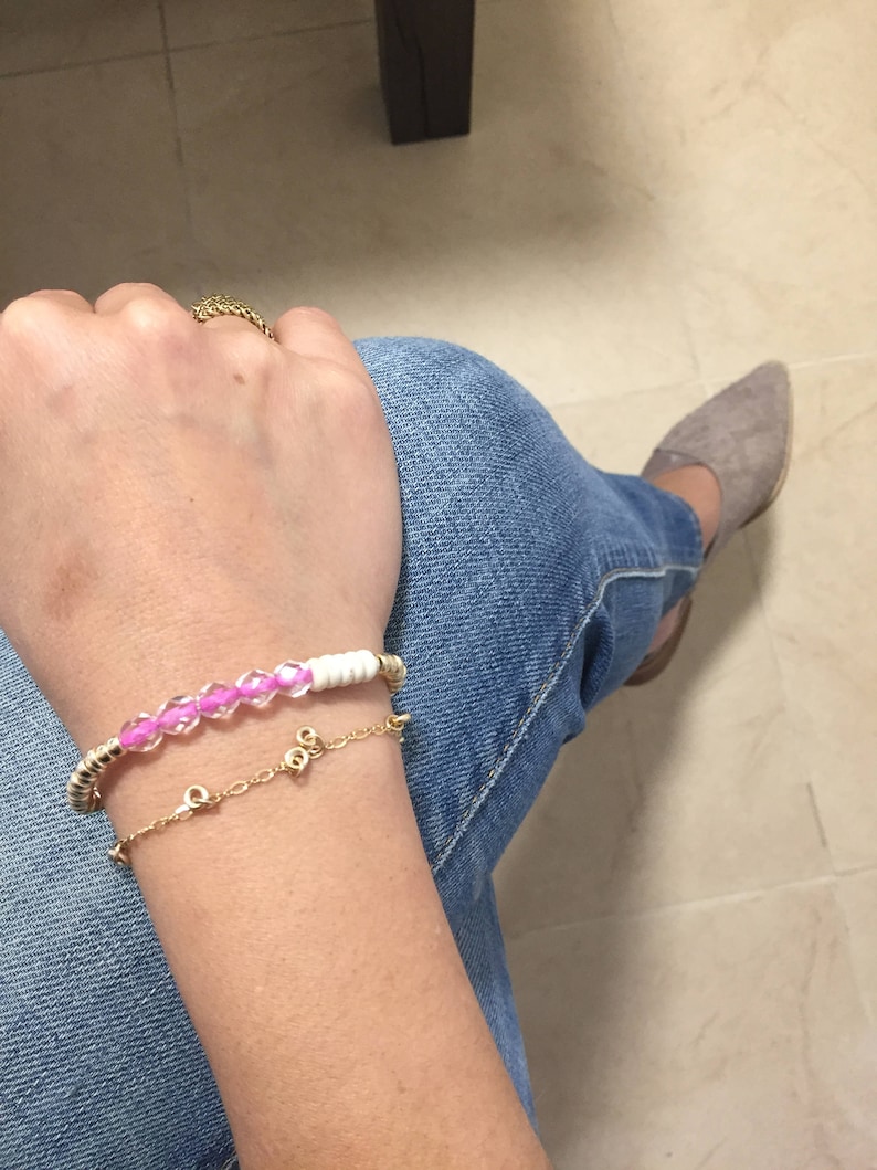 May include: Two bracelets are displayed on a person's wrist. One is a gold chain bracelet with small charms. The other is a beaded bracelet with pink beads and white cylindrical beads, with gold accents. The person is wearing blue jeans.