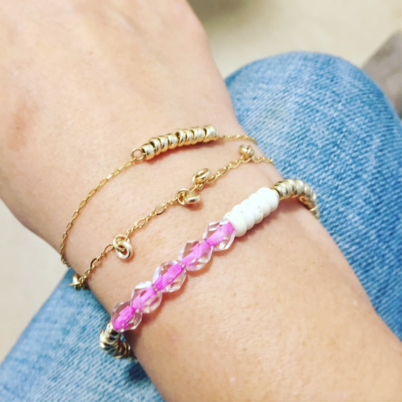 May include: Three bracelets on a wrist. The top bracelet is a delicate gold chain with small gold rings. The middle bracelet is a gold chain with small gold beads. The bottom bracelet has white beads, pink crystals, and gold accents.