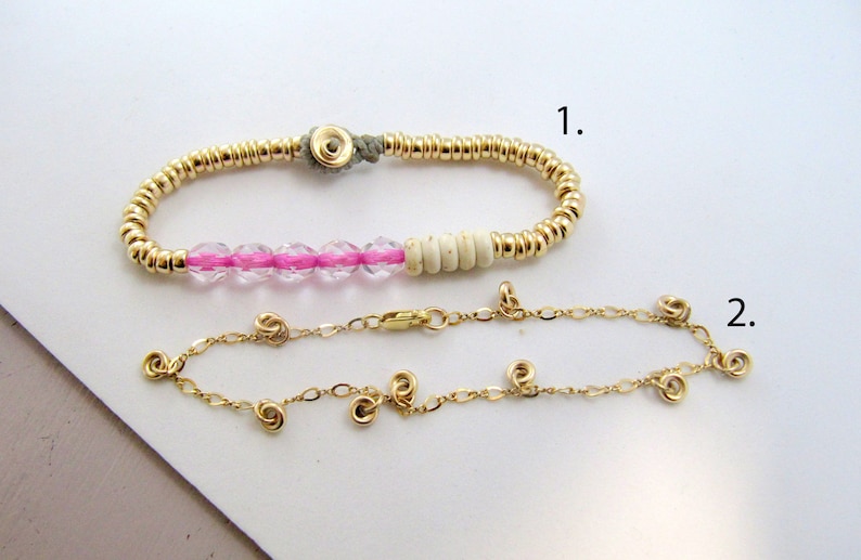 May include: Two bracelets. The top bracelet features gold beads, pink crystal beads, and off-white beads. The bottom bracelet is a delicate gold chain with small, spiral-shaped gold accents. Both bracelets are displayed on a white surface.
