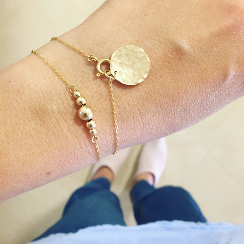 Puede incluir: Dos pulseras doradas se muestran en un brazo. Una tiene peque&ntilde;as cuentas doradas, la otra un disco de oro martillado. Las pulseras son delicadas y elegantes. La joyer&iacute;a es adecuada para el uso diario.
