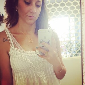 May include: A woman wearing a white lace top with a gold heart necklace and a heart tattoo on her shoulder. She is holding a white iPhone with a green and black phone case that says "Smile".