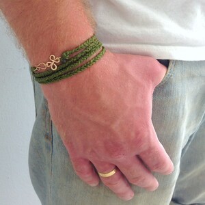 May include: A green braided bracelet with a gold charm on a person's wrist. The bracelet is worn over a white t-shirt and blue jeans.