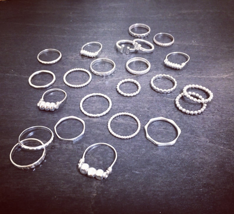 May include: A collection of silver rings, some with textured designs, others with simple bands. The rings are arranged on a dark surface.