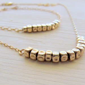 Puede incluir: Un collar de oro con dos filas de peque&ntilde;as cuentas cuadradas texturizadas. El collar est&aacute; sobre una superficie blanca.