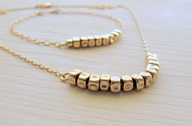 Puede incluir: Un collar de oro con dos filas de peque&ntilde;as cuentas cuadradas texturizadas. El collar est&aacute; sobre una superficie blanca.