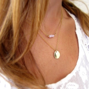May include: A layered gold necklace with a small circle pendant and a delicate chain with tiny pink beads.