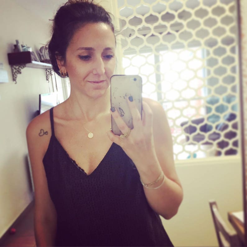 May include: A woman wearing a black lace tank top with a silver necklace and a small silver pendant. She is holding a black cell phone in her right hand and has a small heart tattoo on her left shoulder.
