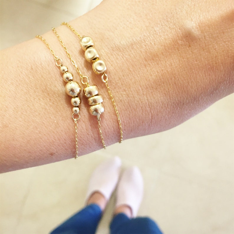 K&ouml;nnte beinhalten: Drei goldene Kettenarmb&auml;nder mit kleinen runden und ovalen Goldperlen. Die Armb&auml;nder sind &uuml;bereinander am Handgelenk einer Person gelegt.