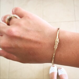Op de afbeelding: Een gouden draad armband met een gedraaid knoop detail. De armband is op een pols.