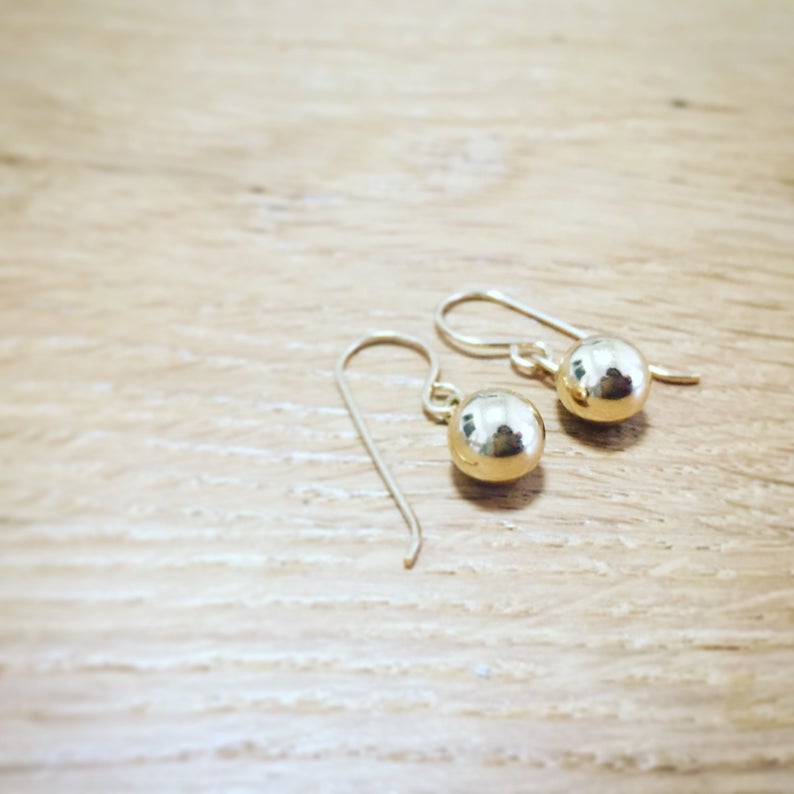 May include: A pair of gold ball drop earrings with a simple design. The earrings are made of gold metal and have a smooth, shiny finish.