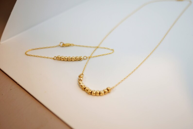 Puede incluir: Un conjunto de collar y pulsera de oro con peque&ntilde;as cuentas redondas. El collar es m&aacute;s largo que la pulsera y las cuentas est&aacute;n dispuestas en forma de curva.