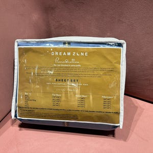 Puede incluir: Un juego de sábanas empaquetado con una etiqueta dorada y blanca. La etiqueta dice "DREAM ZONE Pima Cotton SHEET SET". El embalaje es azul y blanco. El juego de sábanas incluye una sábana encimera, una sábana bajera y fundas de almohada.