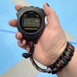 May include: Black stopwatch with the text "STOPWATCH" and a digital display showing "00:00.00". The stopwatch is held by a person wearing a black and rainbow paracord bracelet.