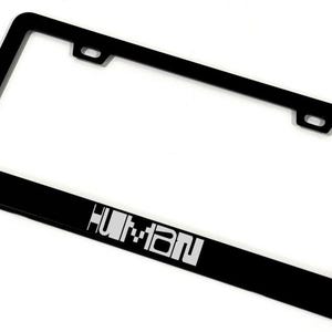 May include: A black license plate frame with the word "HUMAN" in white block letters. The frame has a rectangular shape with rounded corners and two mounting holes at the top.