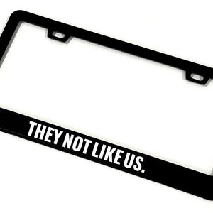 May include: Black license plate frame with the text "THEY NOT LIKE US." The frame has a rectangular shape with rounded corners and mounting holes. The text is in a bold, white font.