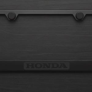 May include: A black license plate frame with the embossed word "HONDA" at the bottom. The frame has a rectangular shape with rounded corners and two circular fasteners at the top. The background is a dark, textured surface.