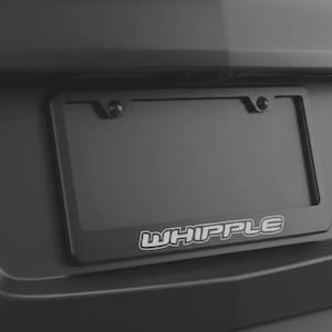 Puede incluir: Un marco negro para matrícula con la palabra "WHIPPLE" en letras blancas. El marco está montado en un vehículo gris oscuro. El marco tiene forma rectangular y está diseñado para una placa de matrícula estándar.
