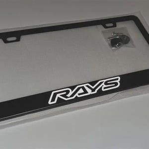 May include: Black license plate frame with the white text "RAYS". The frame is rectangular with rounded corners and includes mounting hardware. The frame is packaged in a clear plastic bag.