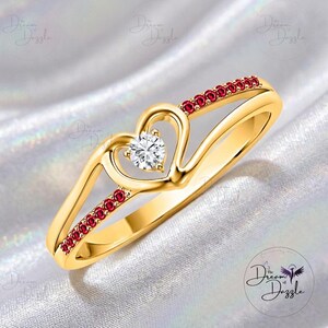 Puede incluir: Un anillo dorado con un diseño en forma de corazón. El anillo presenta una piedra transparente de talla redonda en el centro del corazón y pequeñas piedras rojas a lo largo de la banda. El anillo está sobre un fondo blanco texturizado.