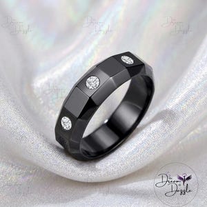 May include: A black ring with a faceted, geometric band design featuring three round, clear, sparkling stones set into the flat surfaces. The ring has a polished, dark finish and a wide, structured profile that highlights its angular, modern shape.