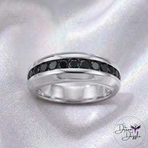 May include: A polished silver band ring featuring a central channel set with a row of round, faceted black stones. The metal has a smooth, reflective finish, and the stones are arranged in a continuous line around the center of the band.