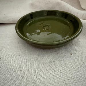 May include: A round, olive green ceramic dish with a glossy finish and a small, embossed leaf design centered at the bottom. The dish features a slightly raised rim and a smooth, reflective surface that catches the light, creating subtle highlights.