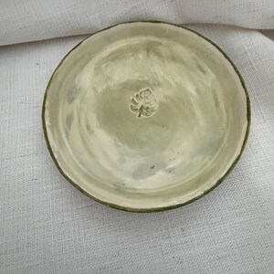 May include: A round, shallow ceramic dish with a glossy, pale sage green glaze. The surface features a subtle, hand-pressed monstera leaf impression centered in the middle. The edges of the dish are slightly uneven, showing a dark green rim.
