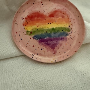 Op de afbeelding: Een rond keramieken bord met een lichtroze basiskleur en een handgeschilderd regenbooghart in het midden. Het oppervlak van het bord is versierd met kleine, veelkleurige verfspatten over het gehele roze achtergrondgebied.