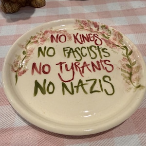 May include: A cream-colored ceramic plate with the words "NO KINGS, NO FASCISTS, NO TYRANTS, NO NAZIS" in red and green lettering. The plate is decorated with a floral border in pink and green.