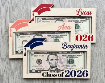Può includere: Tre portadenaro in legno per la laurea con cappelli rossi, corallo e blu. Ogni cornice contiene una banconota da cinque dollari e presenta un nome e il testo "Class of 2026".