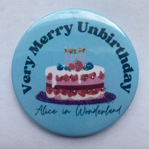 Op de afbeelding: Een ronde, lichtblauwe button met de tekst "Very Merry Unbirthday" rond een taartillustratie. De taart heeft roze lagen, witte glazuur en bessen, met vier kaarsen erop. De tekst "Alice in Wonderland" staat onderaan.