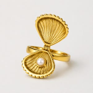 May include: A gold-toned ring featuring a hinged shell design that opens to reveal a single white pearl nestled in the bottom half. The shell has a textured, ribbed surface on both the interior and exterior, with a polished gold band.