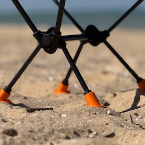 May include: Close-up of a black folding chair on a sandy beach. The chair's legs are black with orange feet. The chair is partially assembled, with the focus on the legs and base. The background shows the ocean.