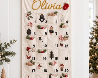 Puede incluir: Un calendario de adviento de tela color crema colgado en la pared, con el nombre Olivia en letras doradas, un pequeño calcetín rojo y veinticuatro bolsillos numerados decorados con motivos navideños y pequeños regalos.