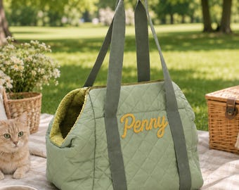 Puede incluir: Un bolso de transporte para mascotas acolchado en color verde salvia con el nombre Penny bordado en letra cursiva amarilla en el lateral. El bolso cuenta con asas largas de tela verde oscuro y un forro interior texturizado verde oliva.