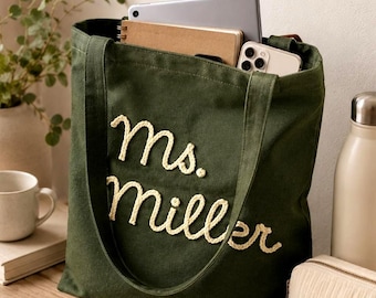 Puede incluir: Un bolso tote de lona verde oscuro con el texto bordado "ms. miller" en hilo color crema claro. El bolso contiene una tableta, un cuaderno y un teléfono inteligente, y está rodeado por una taza, gafas de sol, un estuche pequeño y una cartera.