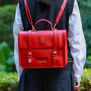 Może przedstawiać: Czerwony plecak w stylu teczki z regulowanymi paskami i górnym uchwytem. Torba posiada złoty emblemat oraz złoty napis "wordswortj Gryffindor". Torba posiada dwa zapięcia na klamry i jest noszona na plecach.