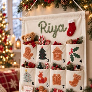 Può includere: Un calendario dell'avvento in tessuto color crema appeso a un'asta di legno, con il nome Riya in filato verde, una piccola calza rossa e otto tasche numerate da 1 a 8, decorate con icone festive come alberi, regali e animali.
