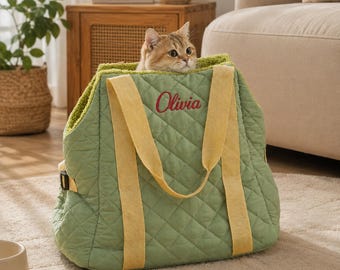 Puede incluir: Un bolso transportín para mascotas de color verde salvia acolchado con asas de tela amarilla y el nombre Olivia bordado en rojo en la parte frontal. Un gato atigrado marrón asoma por la abertura superior del bolso.