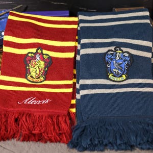 May include: Two knitted scarves, one red and gold, the other blue and gray, with fringed ends. Each scarf features a house crest patch. The red scarf has the name "Alexis" embroidered on it. The scarves are likely themed after a popular fantasy series.