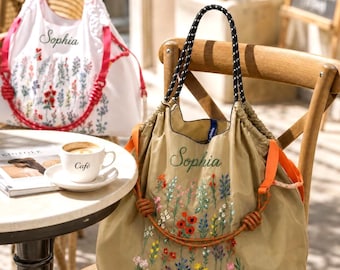 Puede incluir: Un bolso tote de color tostado con el nombre Sophia bordado y un colorido estampado floral. El bolso tiene asas con estampado en blanco y negro y correas laterales naranjas con detalles anudados. Se ve un bolso blanco similar al fondo.